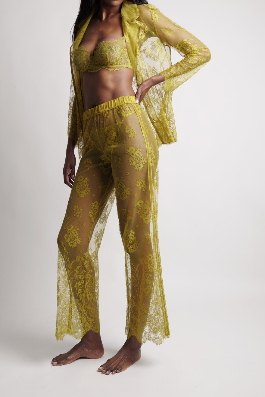 A woman modeling a sheer yellow lace lingerie set with a matching open-front robe, featuring a bralette and wide-leg pants, against a light gray studio background.