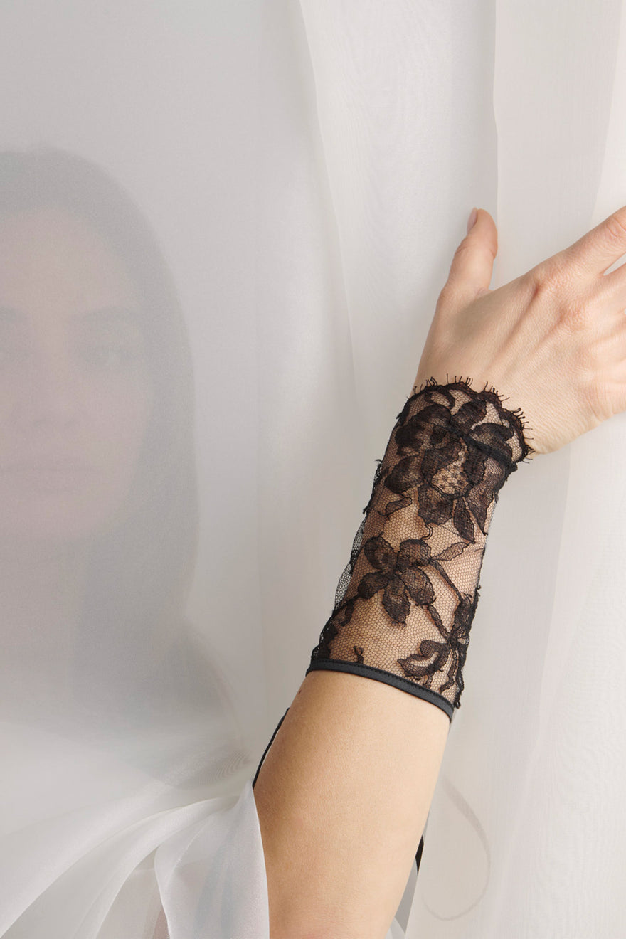 A close-up of a person's hand and forearm wearing a sheer black lace wrist cuff or glove, with a white veil draped in the background.