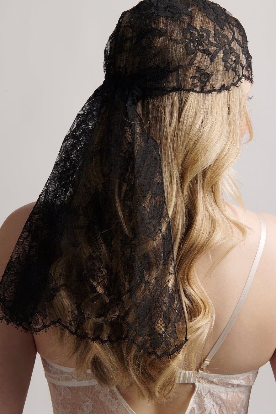 A blonde woman shown from the back wearing a sheer black lace head covering with floral lace detailing and a long drape, paired with a white lace camisole against a light gray studio background.