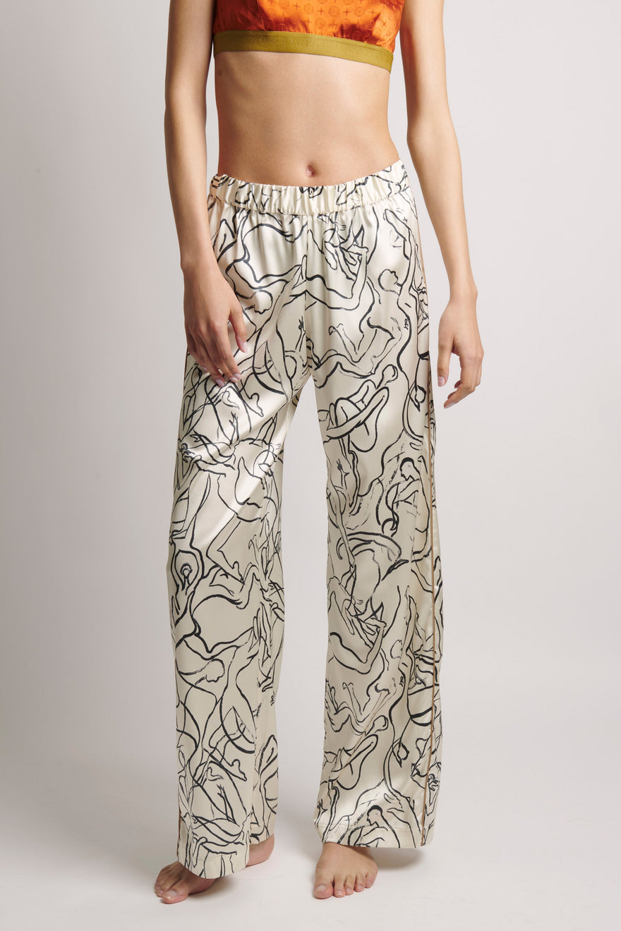 A model wearing high-waisted, wide-leg pants with an abstract black line art print on a cream satin-like fabric, paired with an orange crop top against a light gray studio background.