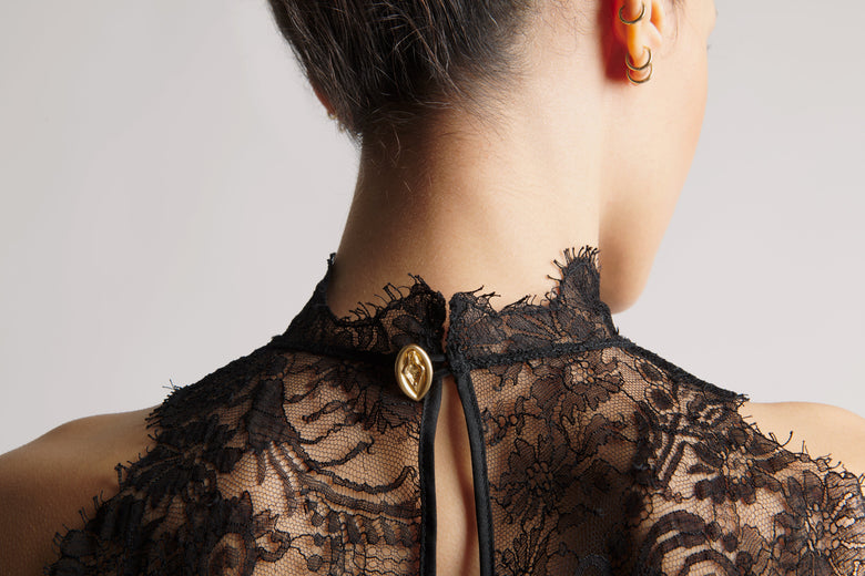 Close-up back view of a person wearing a black lace garment with a high neckline, sheer mesh, and a gold oval button closure at the back.