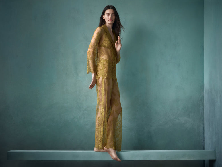 Woman in a golden olive lace matching pajama top and bottom standing on a platform against a teal wall
