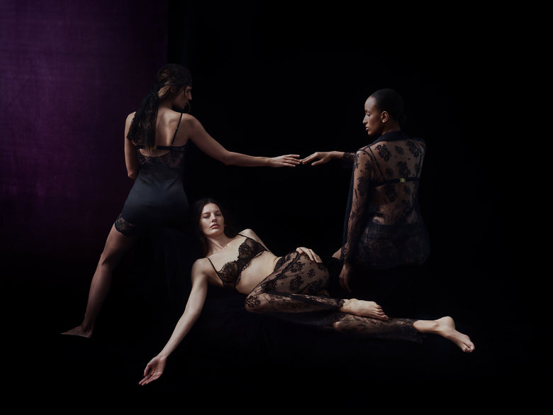 Three models in sheer black lace lingerie and sheer black shapewear pose against a dark studio background with purple lighting on the left.