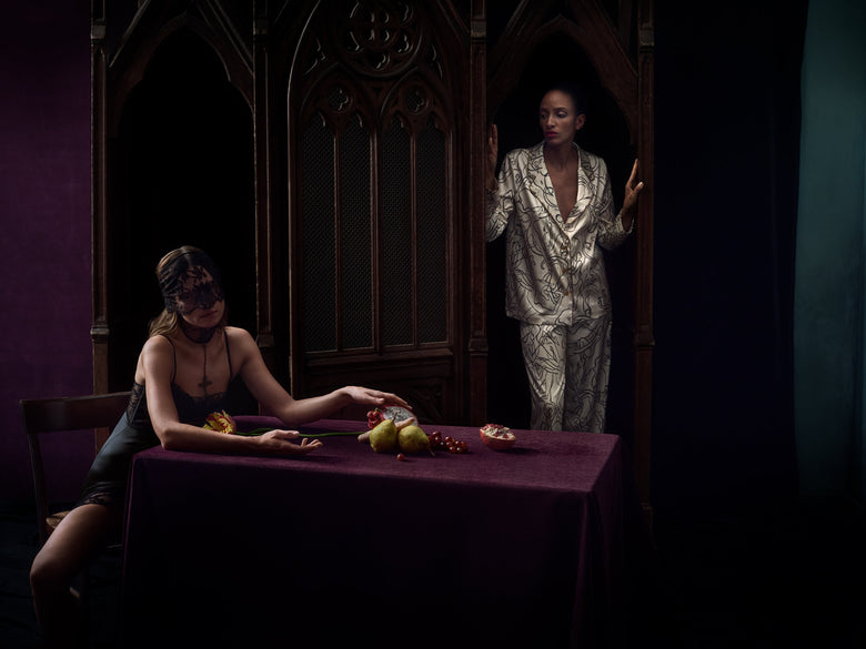 A moody fashion photograph featuring two models in a gothic-themed room. One model is seated at a table draped in a deep purple cloth, wearing a black lace veil and a black dress, while the other stands in a doorway wearing a shimmering silver patterned robe.