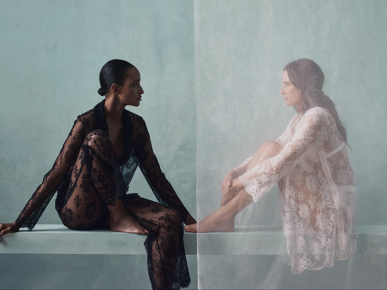 Two women in sheer lace garments pose on a low platform against a pale blue wall and translucent drape. One wears a black lace robe and matching pants, seated in profile; the other wears a white lace robe and sits slightly higher, with one foot extended toward the first woman.