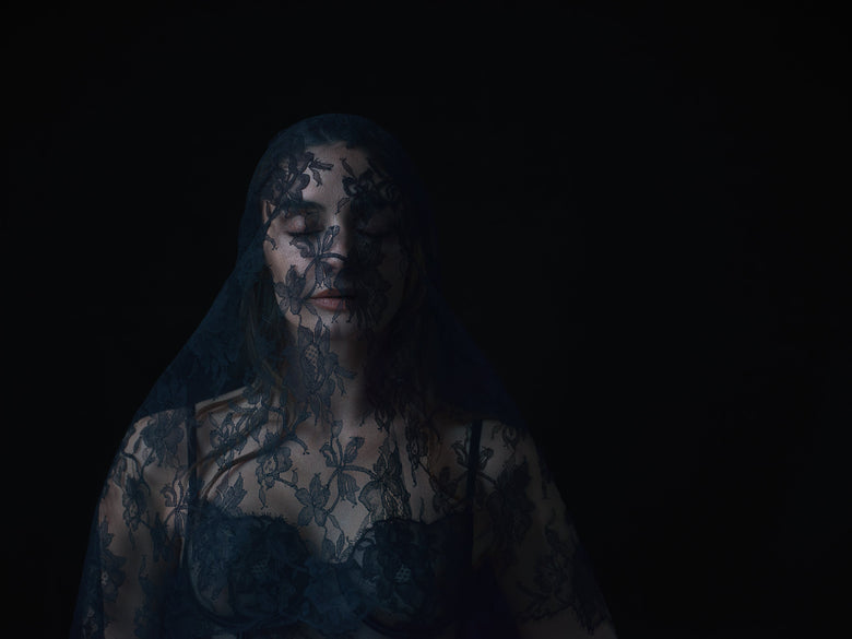 A woman wearing sheer black lace lingerie and a matching lace veil, posed against a solid black background.