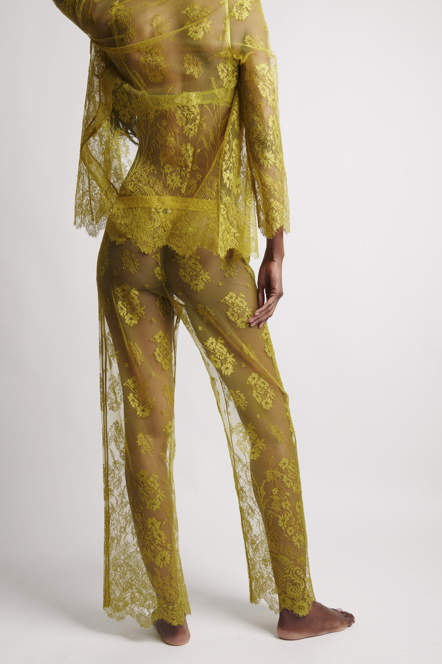 A model is shown from the back wearing a sheer olive green lace pajama set with matching long-sleeve top and pants, standing barefoot against a light gray studio background.