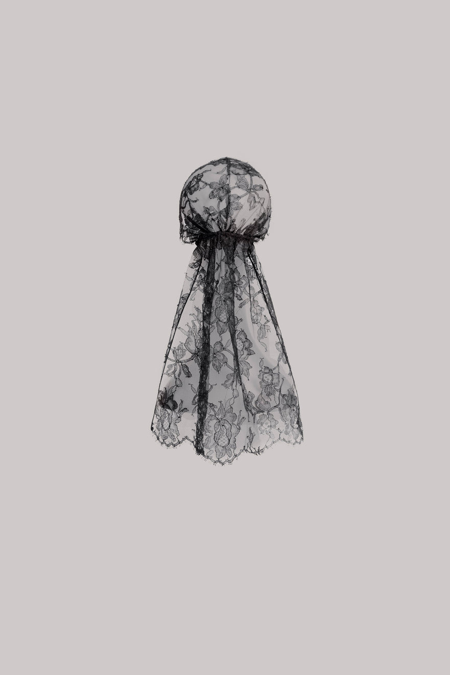 A black lace head covering or veil with a sheer floral pattern and scalloped edges, shown against a solid light gray background.