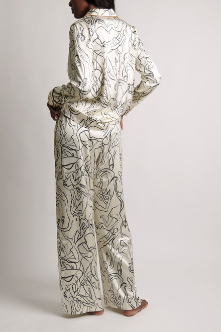A model shown from the back wearing a matching long-sleeve collared shirt and wide-leg pants set in a shiny cream satin fabric with a black abstract line pattern, set against a plain light gray studio background.