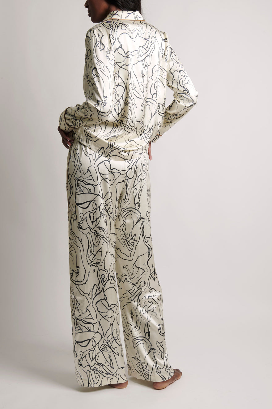 A model shown from the back wearing a matching long-sleeve collared shirt and wide-leg pants set in a shiny cream satin fabric with a black abstract line pattern, set against a plain light gray studio background.