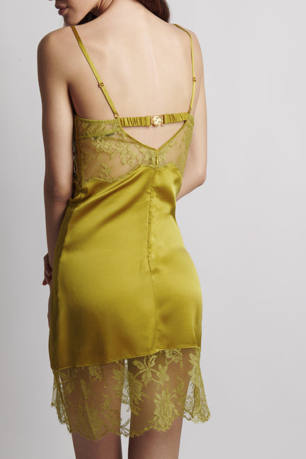 A woman shown from the back wearing a satin slip dress in a muted olive yellow-green color with matching floral lace trim and thin straps.