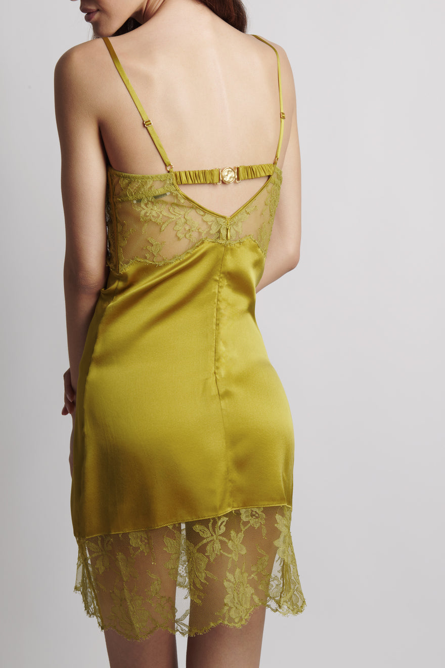 A woman shown from the back wearing a satin slip dress in a muted olive yellow-green color with matching floral lace trim and thin straps.