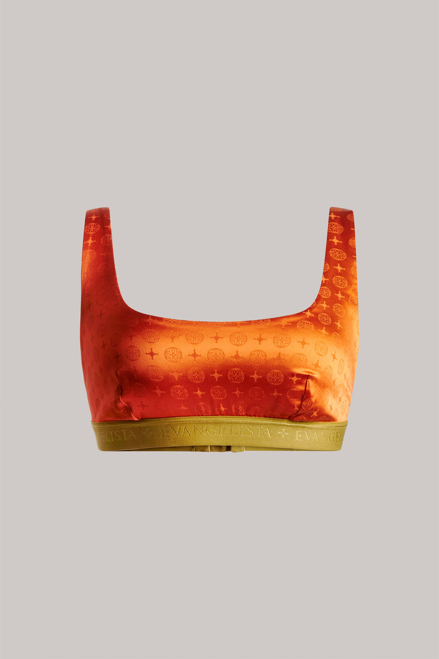 A cropped silk sports bra in a vibrant orange and red gradient with a subtle monogram pattern, featuring a solid olive green underband, shown against a solid gray background.