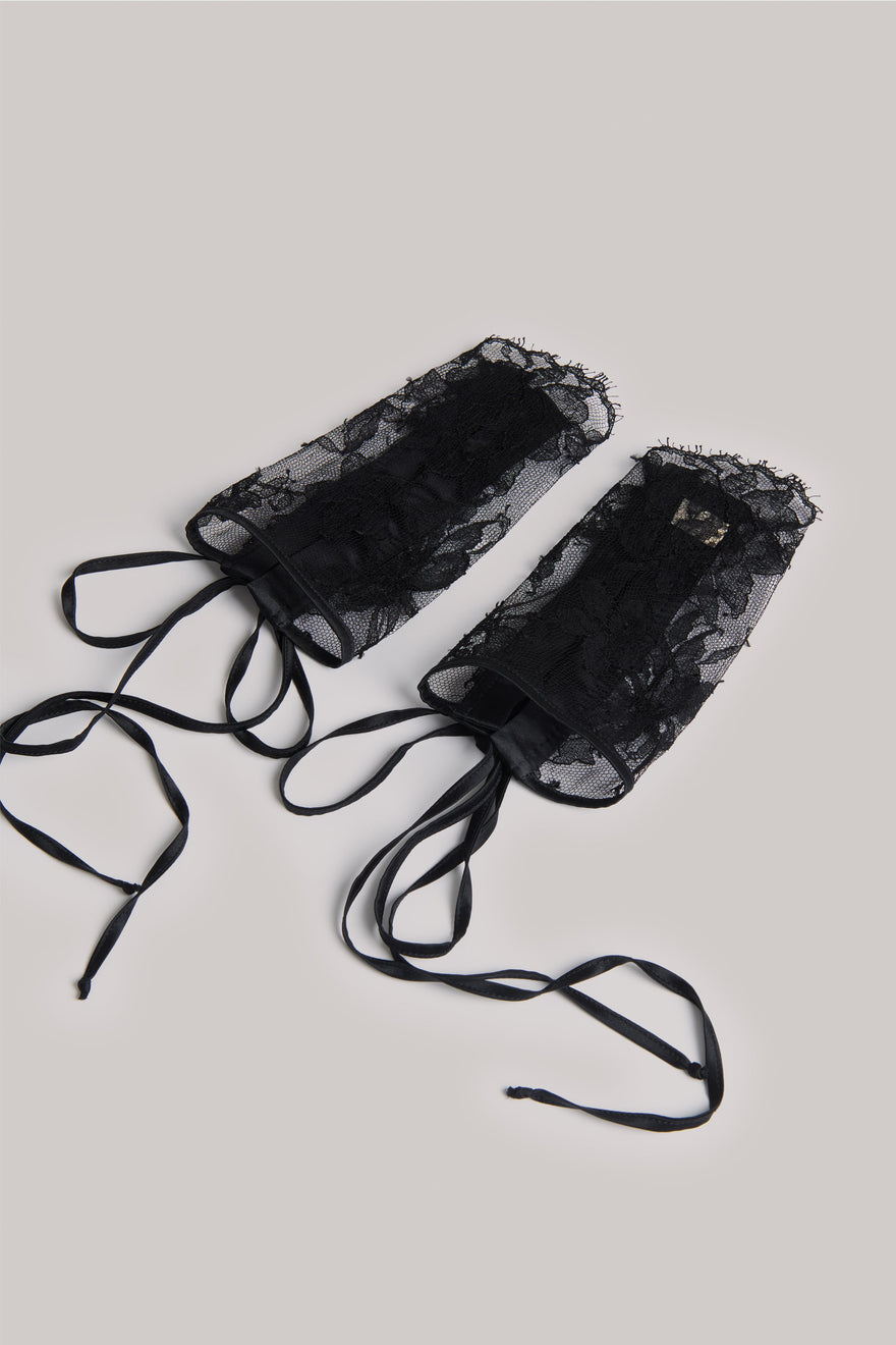 A pair of black sheer lace wrist cuffs with ruffled trim and long tie straps, laid flat on a light gray background.