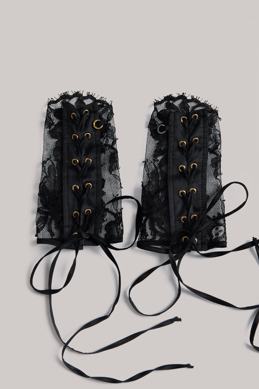 A pair of black lace corset-style waist ties with gold-tone eyelets and long black laces, laid flat on a light gray background.