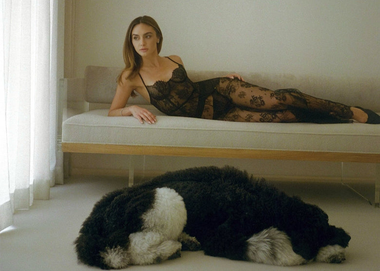 The founder, Christine Evangelista reclines on a low bed wearing a sheer black floral lace bodysuit and matching lace tights, while a large black-and-white dog lies curled up on the floor in the foreground of a softly lit room.